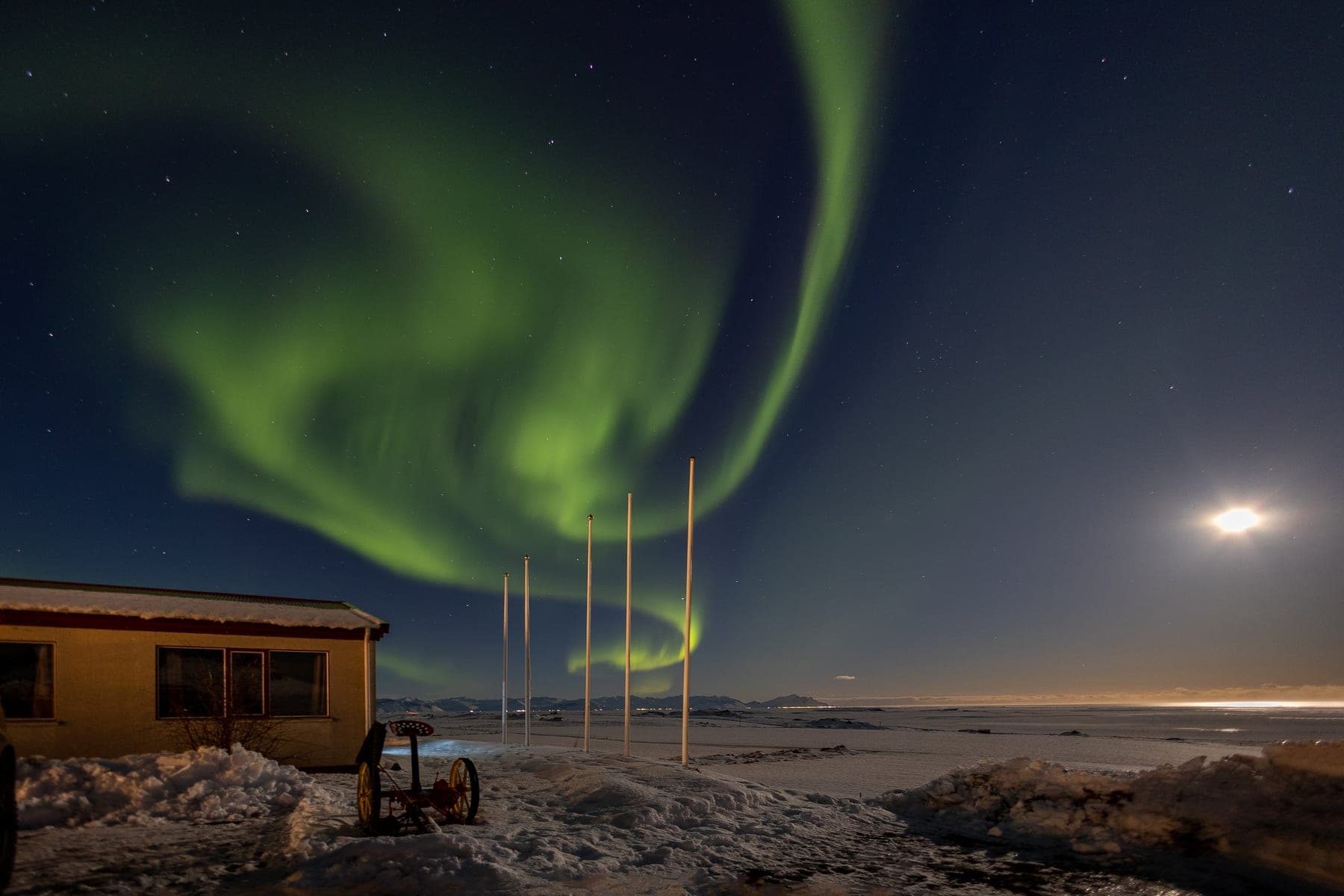 Green aurora borealis curling across the night sky above a lit cabin with the moon low on the horizon