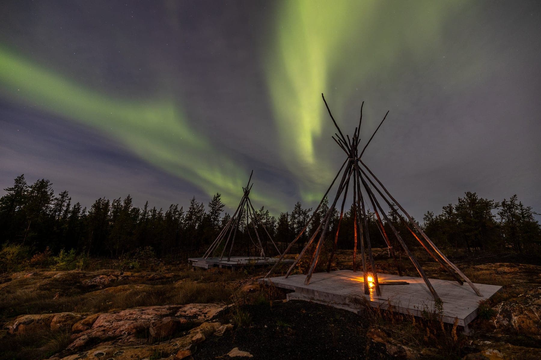 Green aurora arcing over wooden teepee frames on a rocky northern shoreline at night
